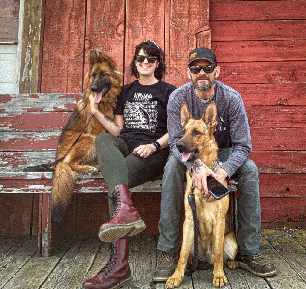 Man and woman sitting on a bench with two German Shepherd dogs.