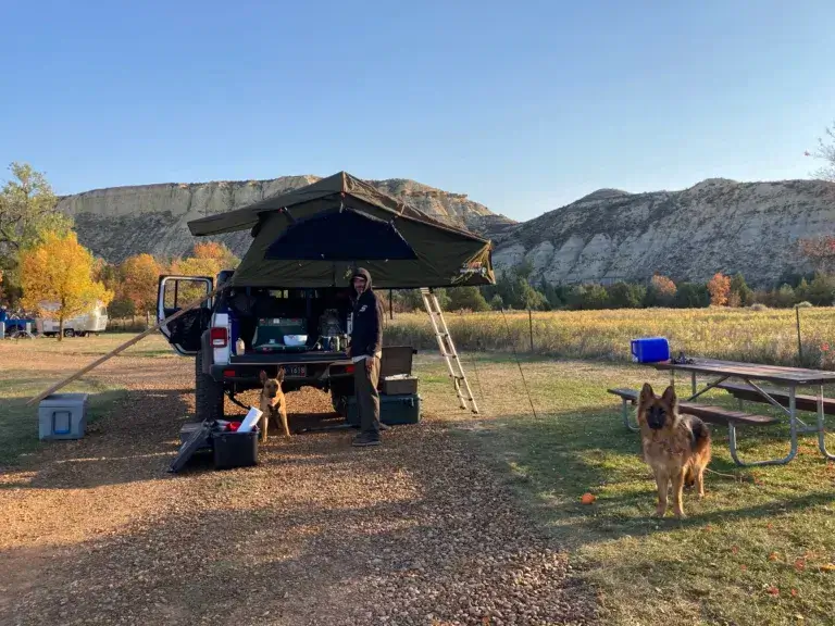 A man with two German Shepherd dogs standing by a Jeep Gladiator with a rooftop tent in a campground near Badlands National Park.