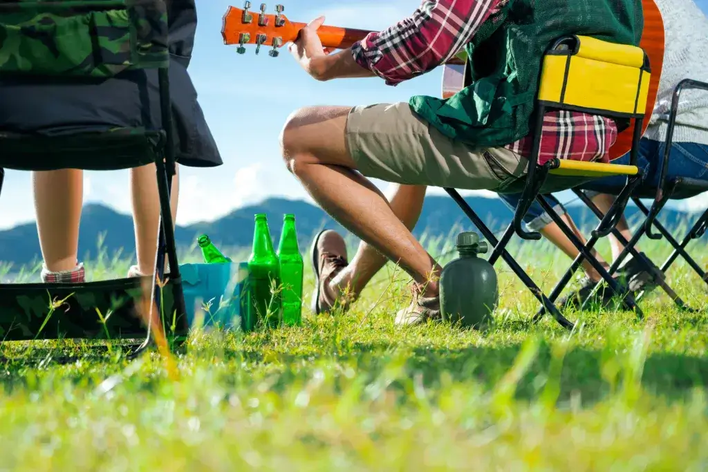 A cropped photo of group of Asian friends sitting on chairs, singing and drinking some beer and water together outside the tent while they are camping.