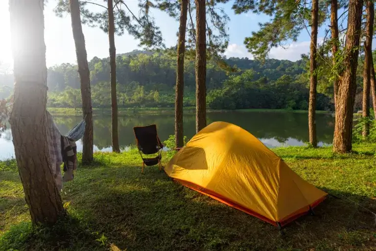 Orange tent next to a lake with a hammock tied between two trees and a camp chair.
