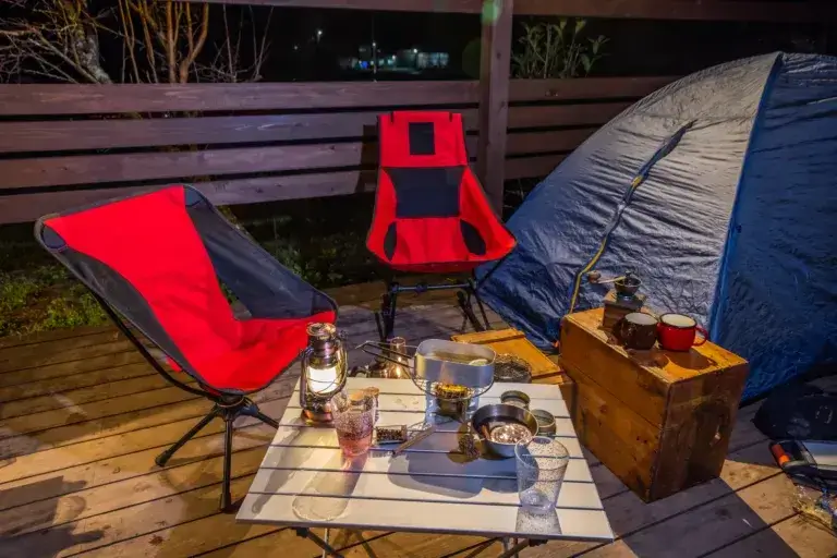 Two red camping chairs next to a tent and a table with lanterns and food.