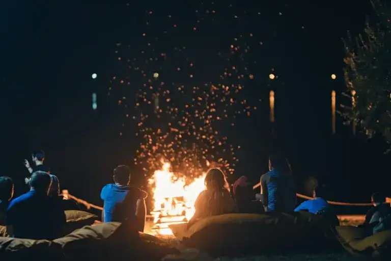 Group of people near camp fire with campfire song and campfire meals playing campfire games and eating campfire grill, telling campfire stories near the fire with wood, flames in the nature at night.