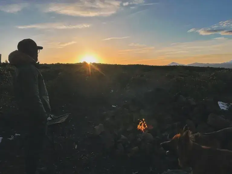 Man and two dogs standing next to a campfire at dusk with mountains in the background.