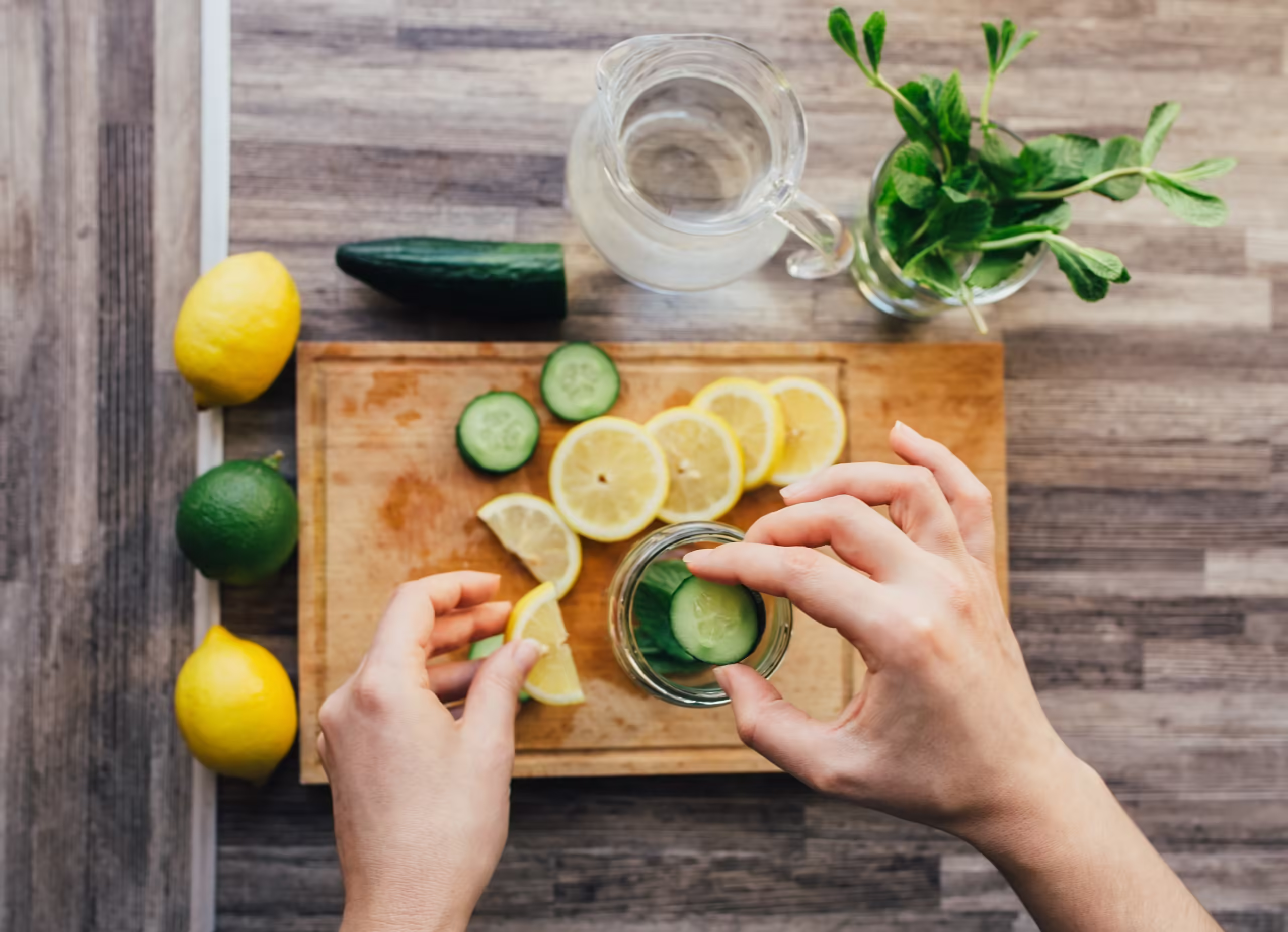 Women's hands preparing detox water with lemon, lime, mint and cucumber. Ingredients for infused water on a wooden board. Top view. Detox, diet concept. Healthy drink