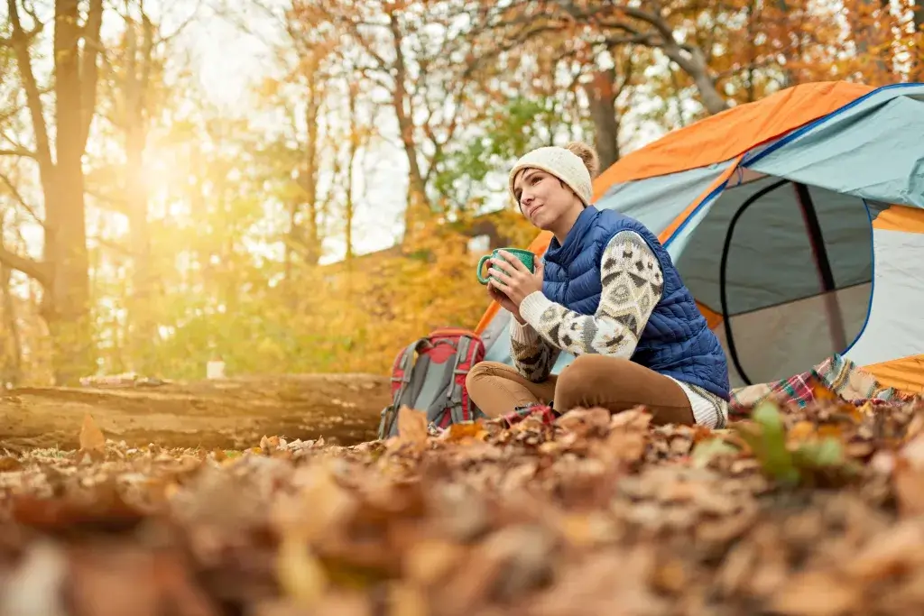 Woman sitting in fall leaves next to a tent drinking a warm beverage.