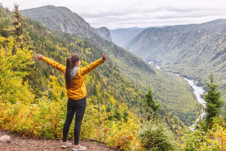 Happy woman hiking up mountain enjoying nature landscape river view from mountain top trail hike. Girl with open arms outstretched in joy enjoying fall autumn Jacques Cartier, Quebec, Canada travel.