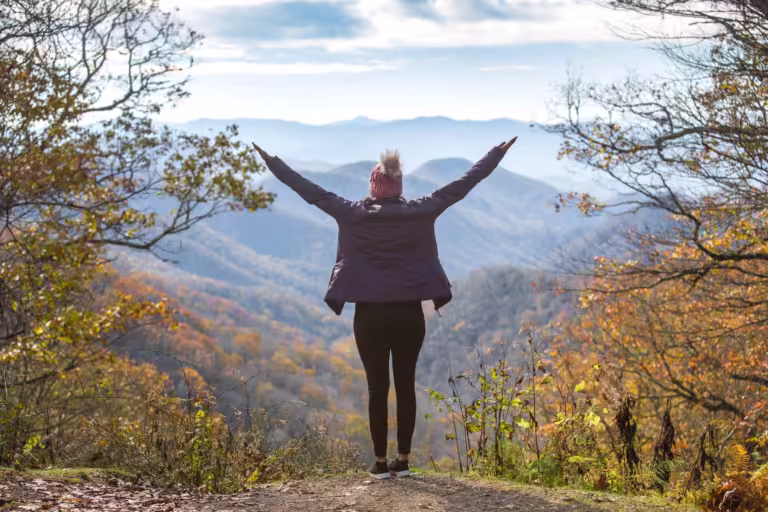 A young woman looks out over the Smoky Mountains in Autumn as she raises her hands to the sky spiritually