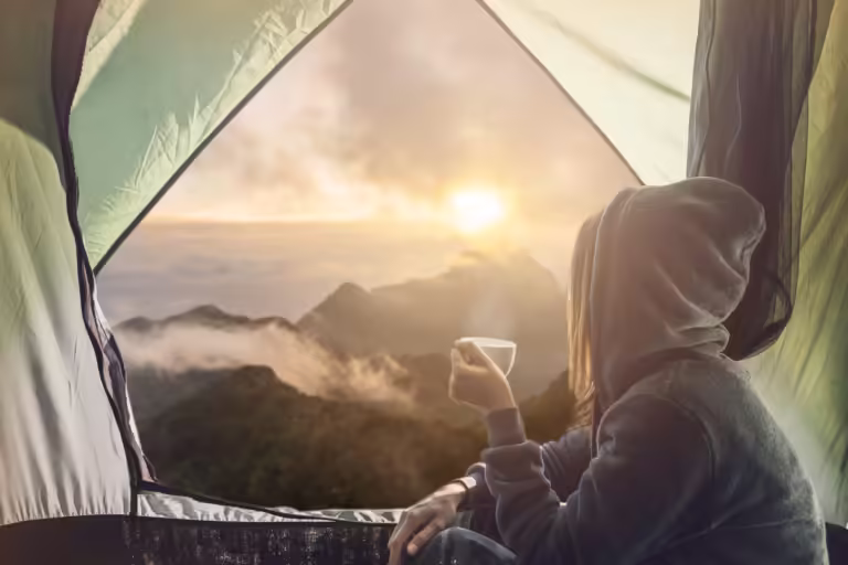 Young woman traveler sitting in the tent, relaxing and drinking coffee with beautiful sunrise.
