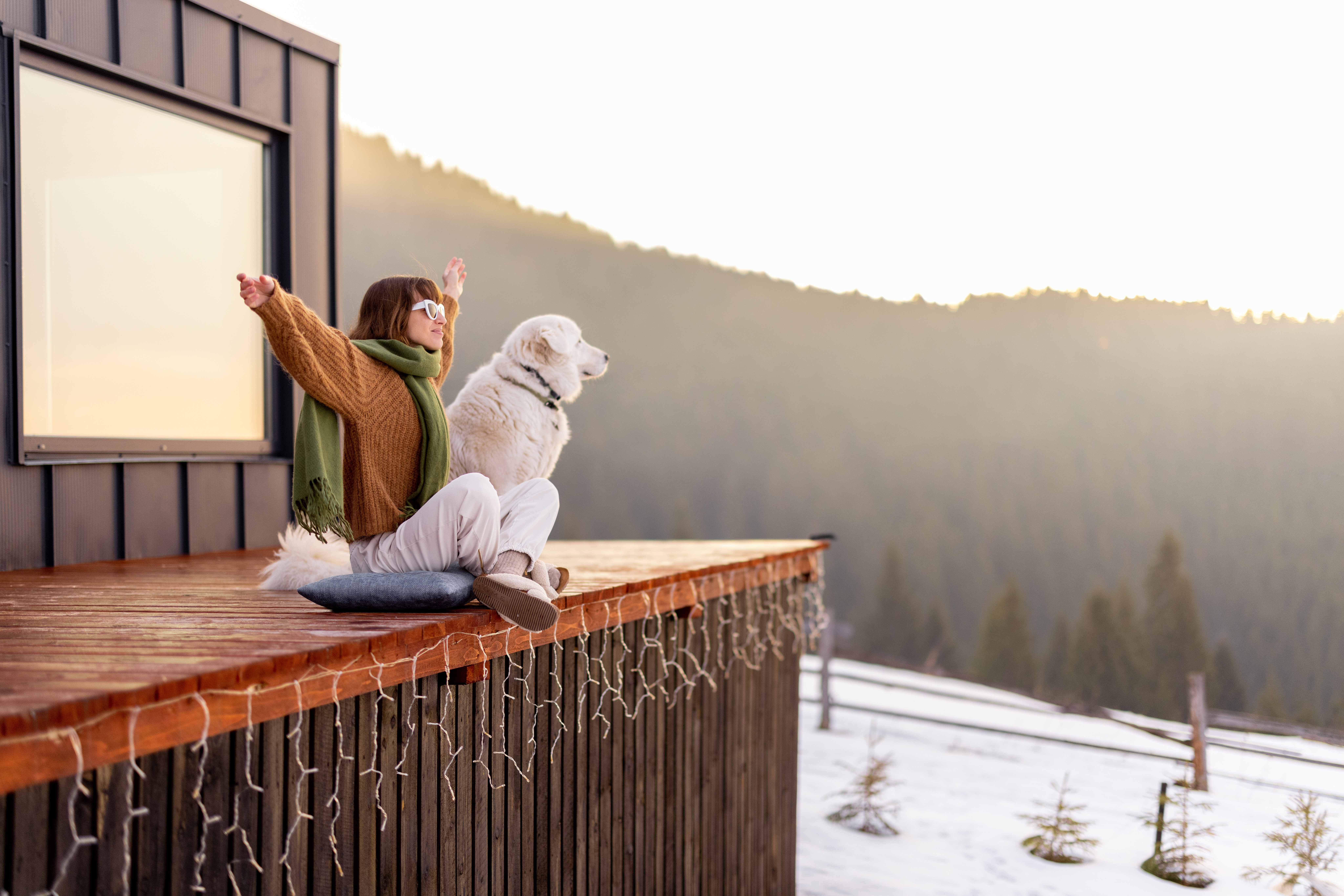 Woman sitting with dog on terrace of tiny house in the mountains enjoying beautiful sunrise landscape. Concept of small modern cabins for rest and escape to nature. Idea of traveling with dog