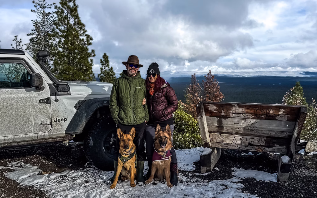 Man, woman and two German Shepherds standing next to a Jeep with beautiful mountains in the background.