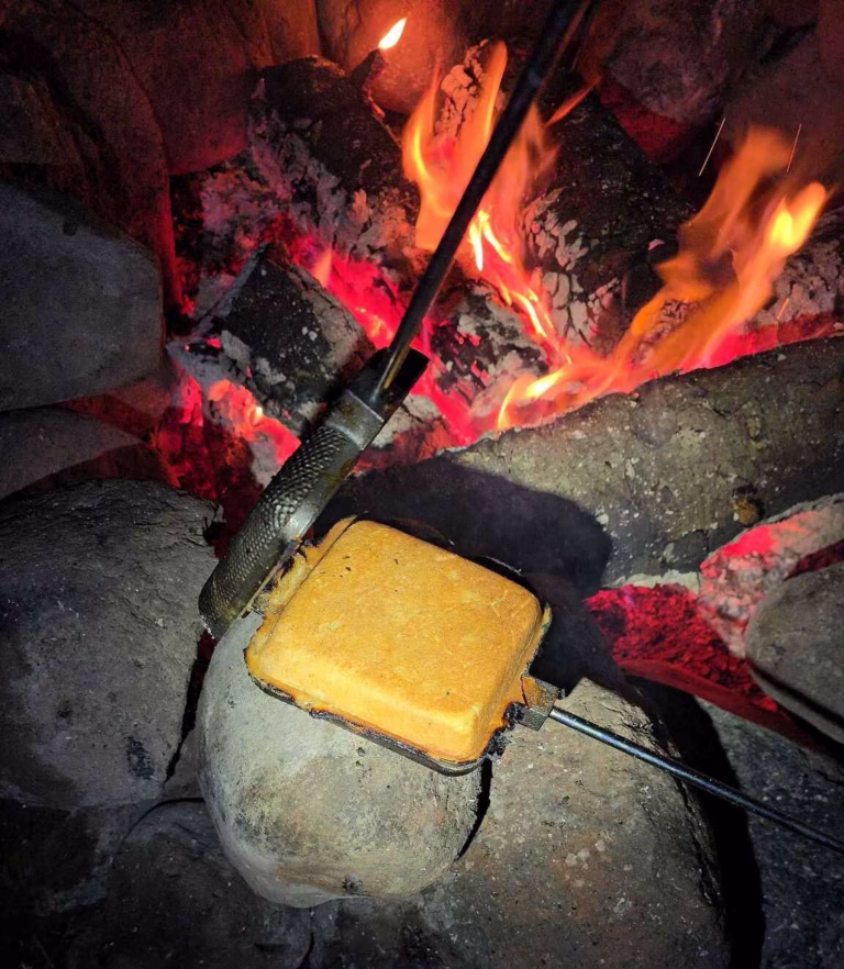Opening cast iron pie iron to reveal a golden brown pie over coals.