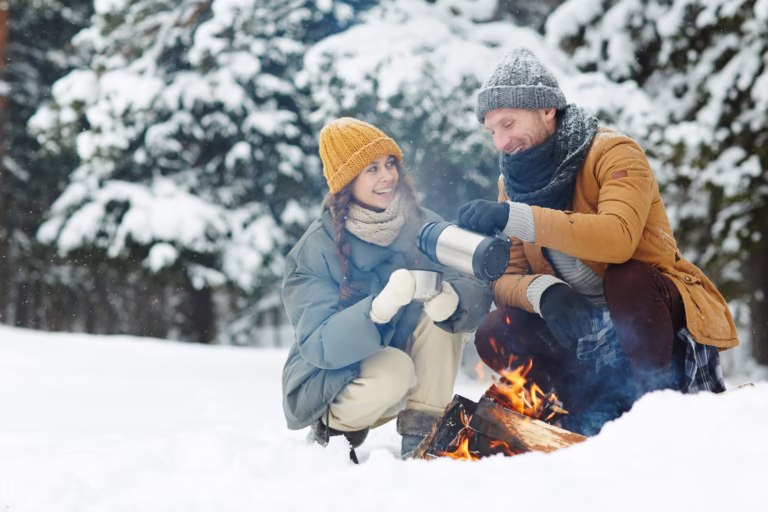 Happy excited young couple in warm jackets drinking hot tea and sitting by fire while enjoying camping in winter forest