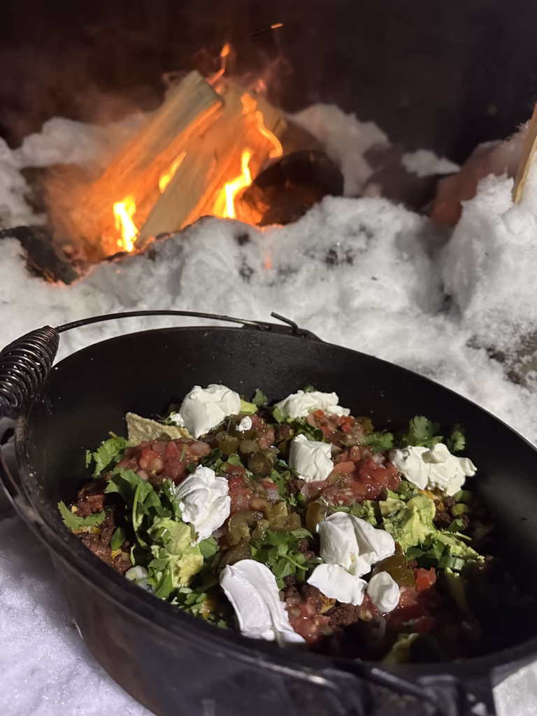 Dutch oven sitting next to a campfire with the contents for nachos.