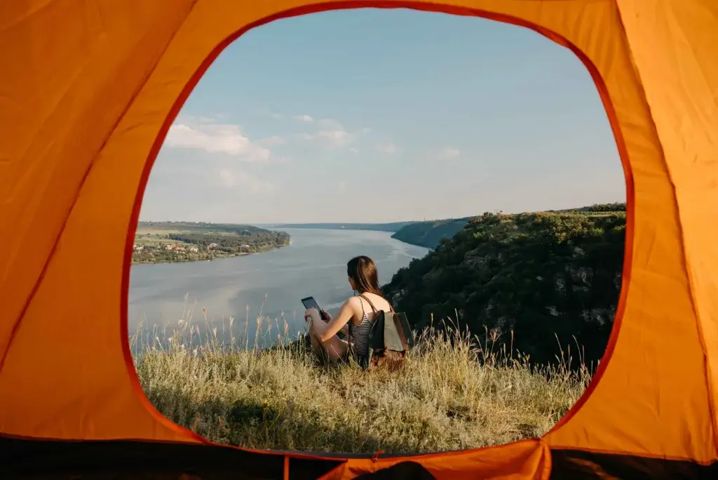 View from a tent over a river with a young womah sitting in the grass looking at a phone.