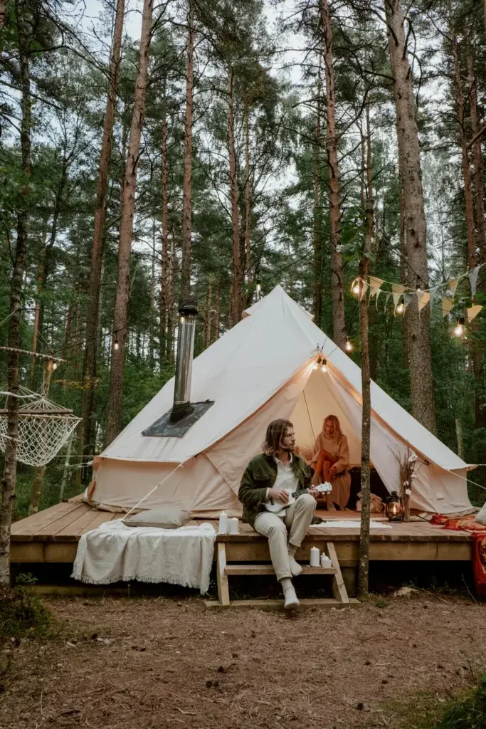 A couple enjoying a cozy glamping experience in a forest with a tent and musical instrument.