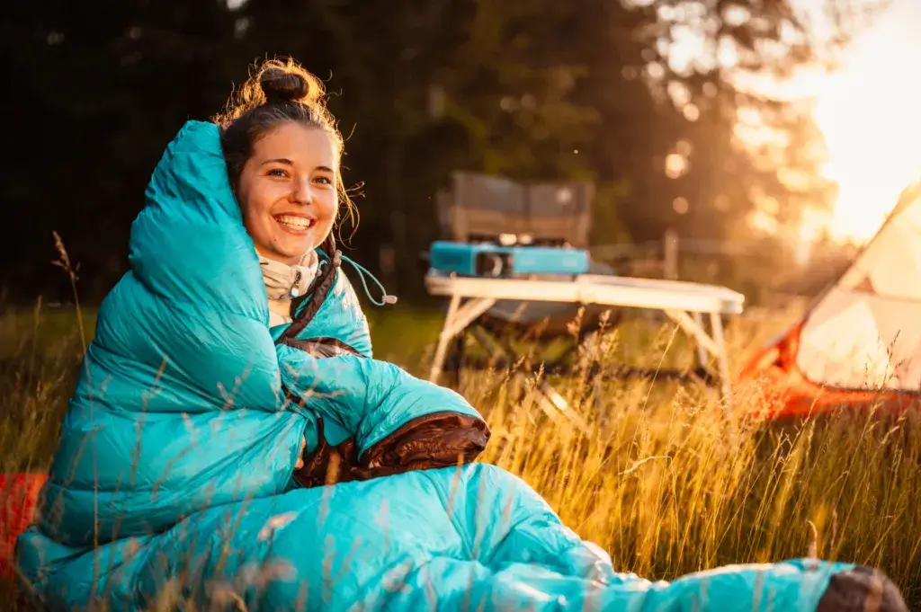 Young woman wrapped up in a blue sleeping bag with a camp stove in the background.
