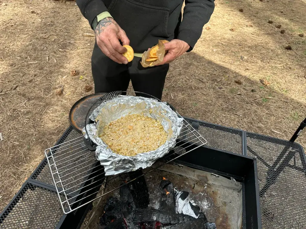 Dutch oven ranch mac n cheese over a campfire with a man putting crushed Ritz Crackers on the top.