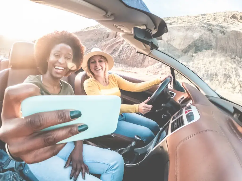 Happy girls having fun in convertible car in summer vacation - Young women friends taking selfie on cabriolet auto outdoor - Main focus on blond girl face - Travel, holidays and friendship concept