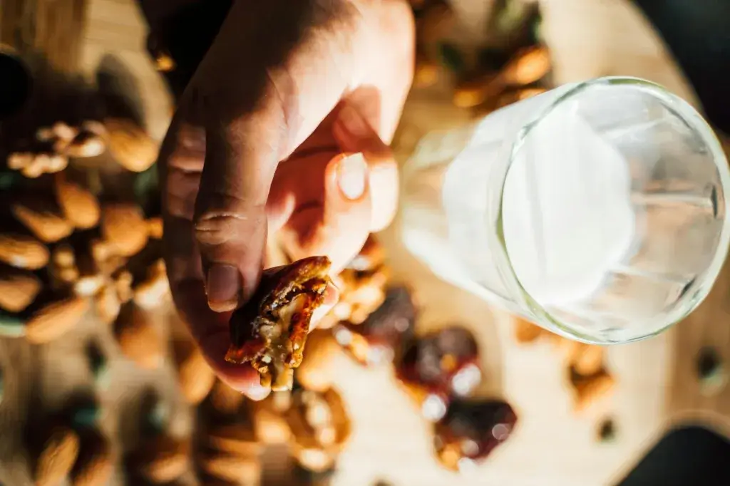 Close-up of a hand holding a date with nuts, surrounded by almonds and walnuts, next to a glass of milk.