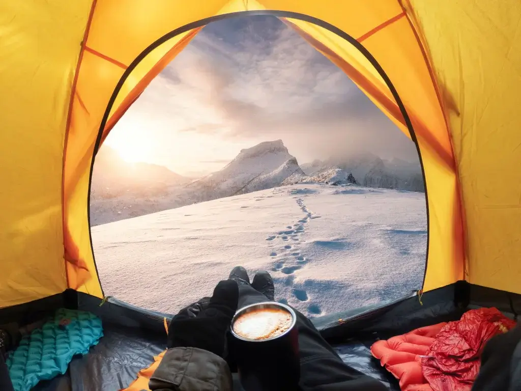 Person lying in a tent holding a cup of hot coco looking outside at the snow.