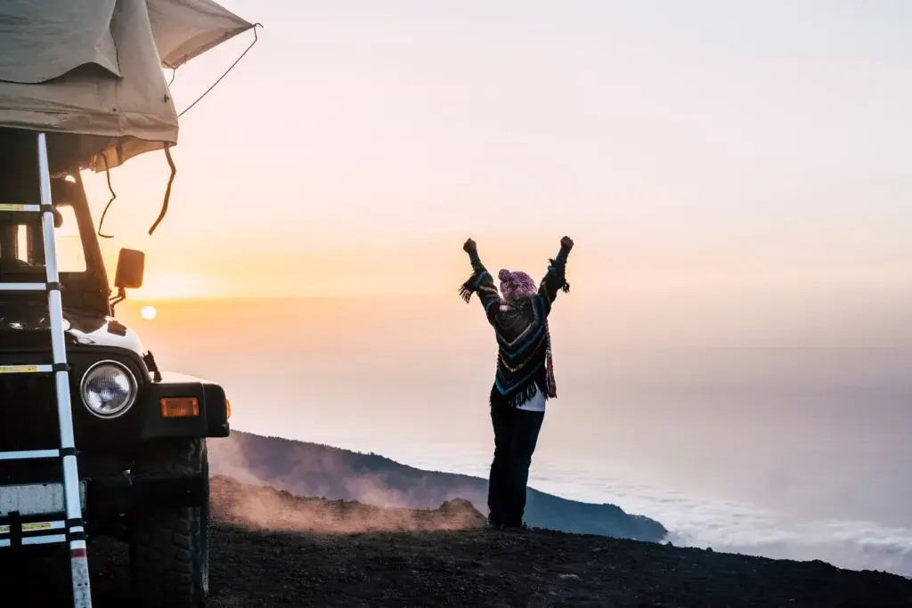 Woman solo camping in a Jeep with a rooftop tent raising arms to the sky at sunset.