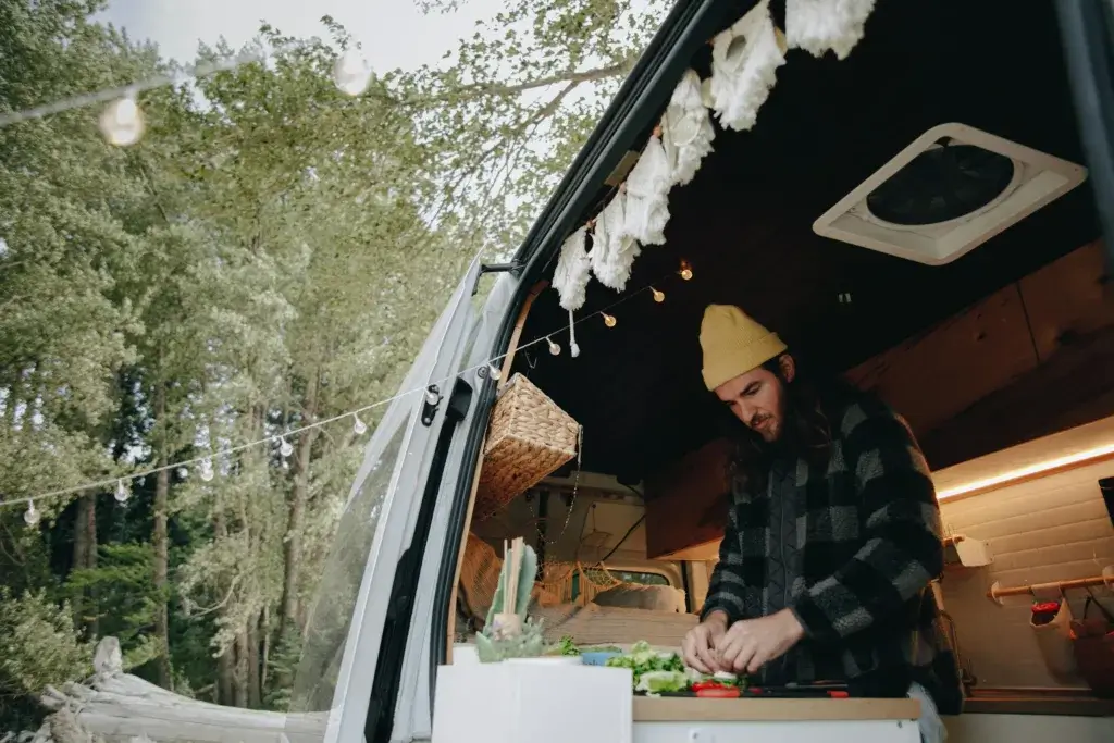 A man in a camper van kitchen prepares food amidst a scenic forest setting, showcasing van life and outdoor adventure.