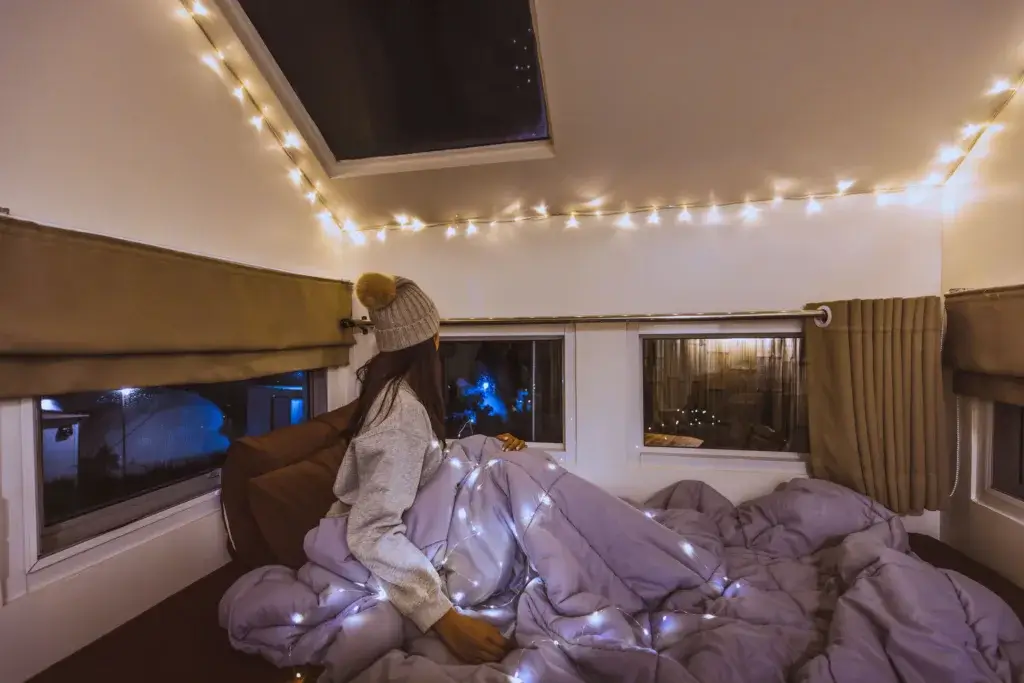 Women in bed in an RV with a warm blanket and hat and string lights.
