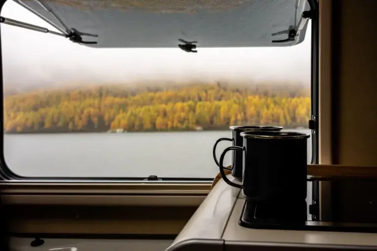 Two mugs sitting on a table in a RV with autumn colors outside the window.