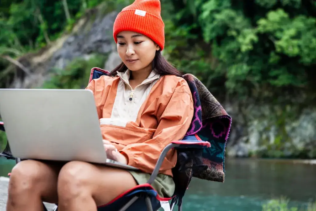 Woman sitting in a camp chair next to a creek working remotely on a laptop.