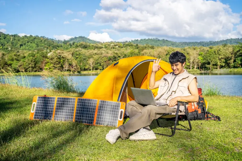 Young man sitting at a camp chair with a laptop and solar setup working off grid from a tent in the background.