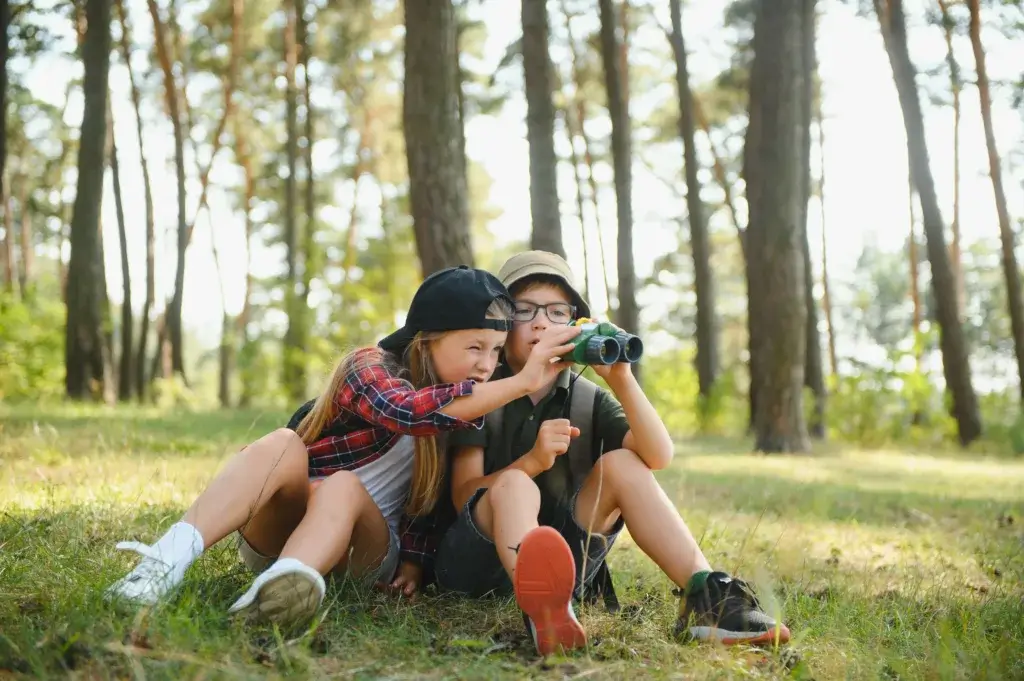 Two children are sitting in the grass with a pair of binoculars.
