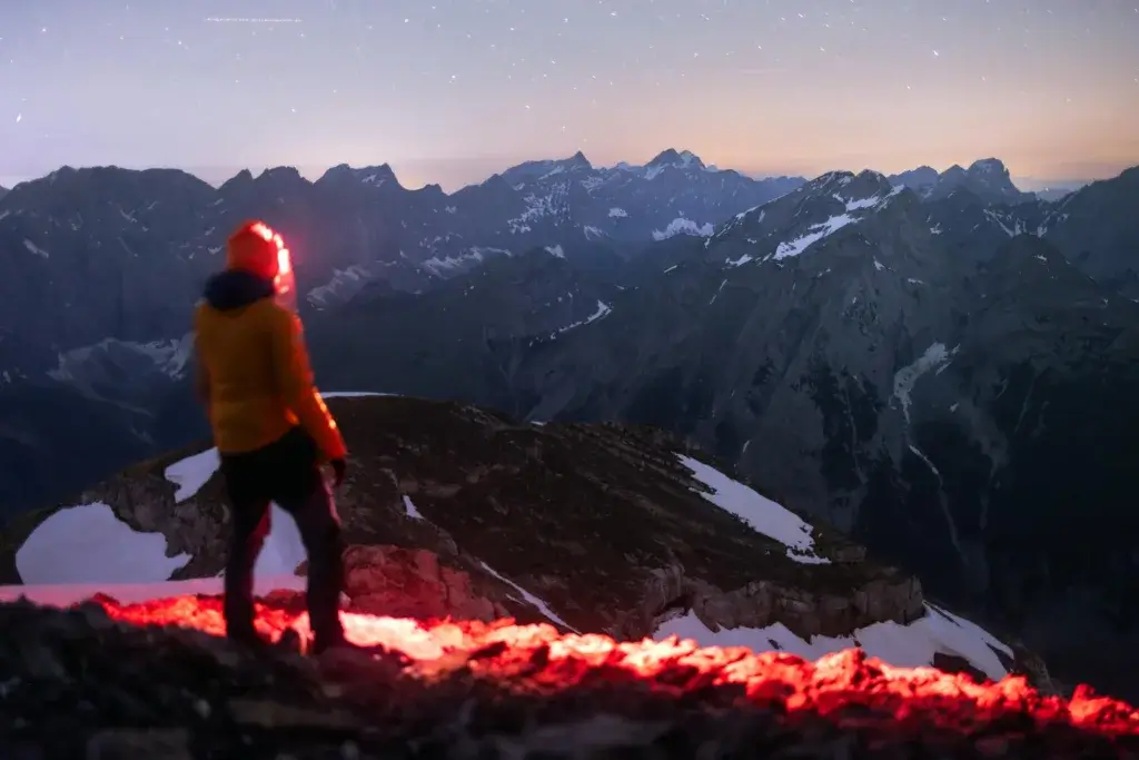 A lone hiker with a headlamp stands on a mountain peak under a starry Alpine night sky.