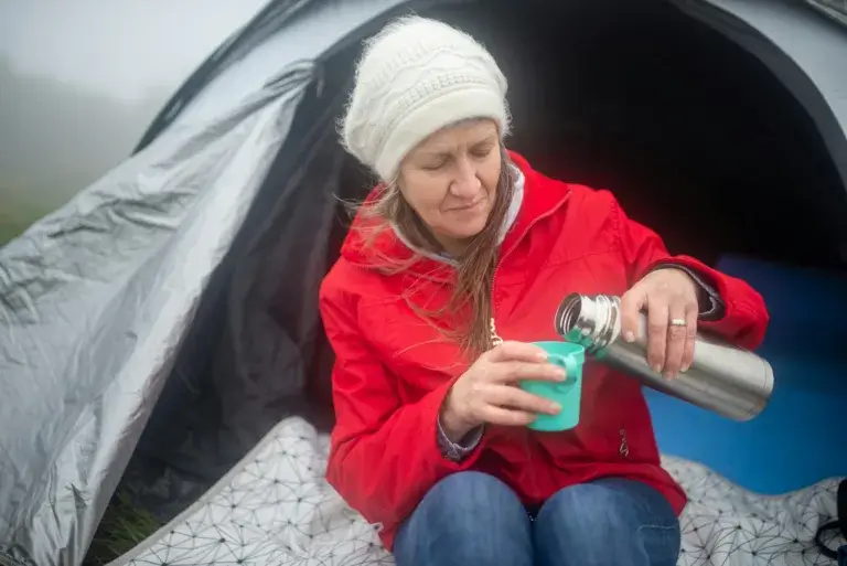Caucasian woman in a red jacket camping, pouring a drink from a thermos by her tent in Portugal.