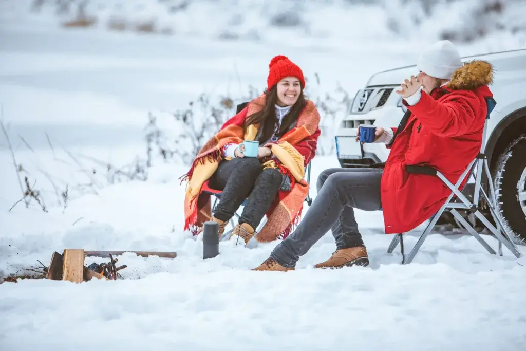 Two women siting next to a campfire drinking warm drinks and car camping.