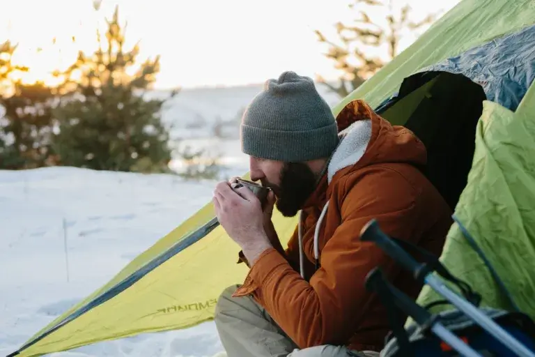 A man is drinking a warm drink in a tent in the snow.