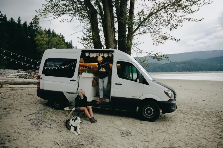 A couple with their dog relax by a camper van on a serene beach, embracing outdoor lifestyle.