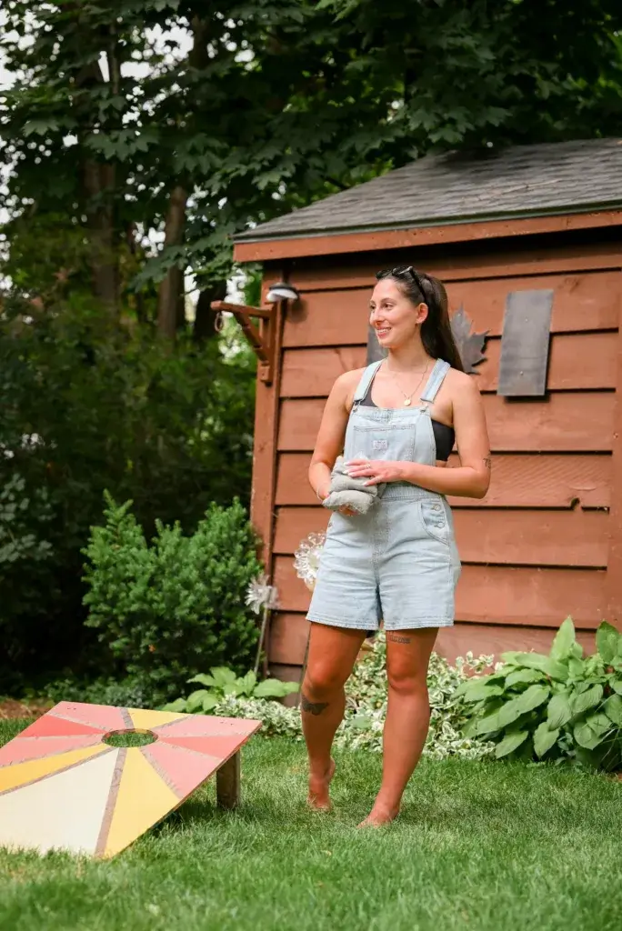 A woman enjoys a game of cornhole in a summer backyard, dressed casually.
