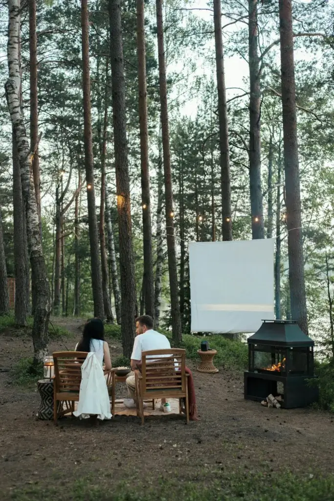 Couple sitting in a forest enjoying an outdoor movie night by a cozy fireplace.