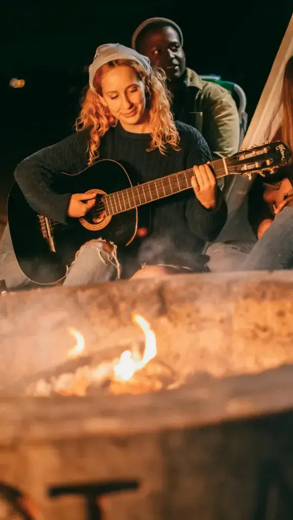 Group of friends playing guitar and singing by a campfire, creating a warm and joyful atmosphere at night.