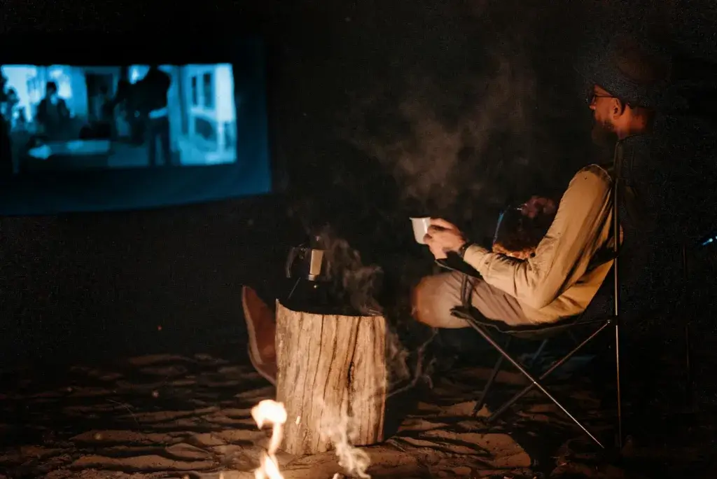 Man enjoying a night of relaxation, watching a movie by a campfire outdoors.