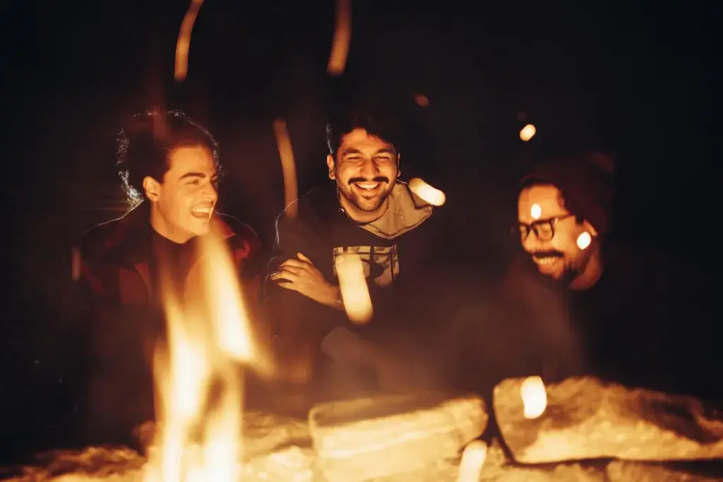 Three friends enjoying a warm campfire at night, sharing smiles and laughter.