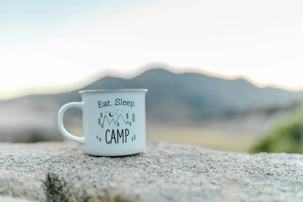 White camping mug with 'Eat. Sleep. Camp' outdoors on a rock.