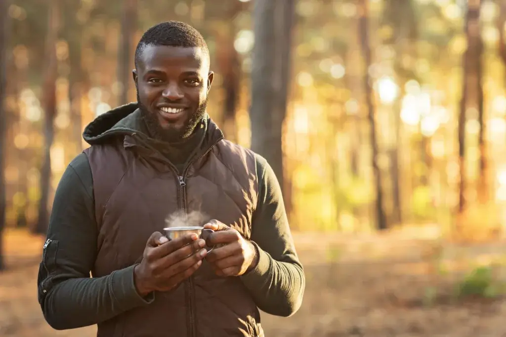 A man is smiling and drinking a hot cup of coffee in the woods.