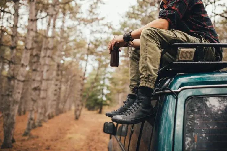A man dressed in outdoor gear is sitting on a car roof in the woods.