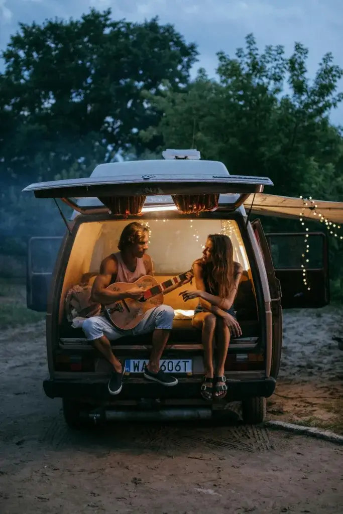 Two adults enjoying a musical road trip evening in a camper van, surrounded by nature.