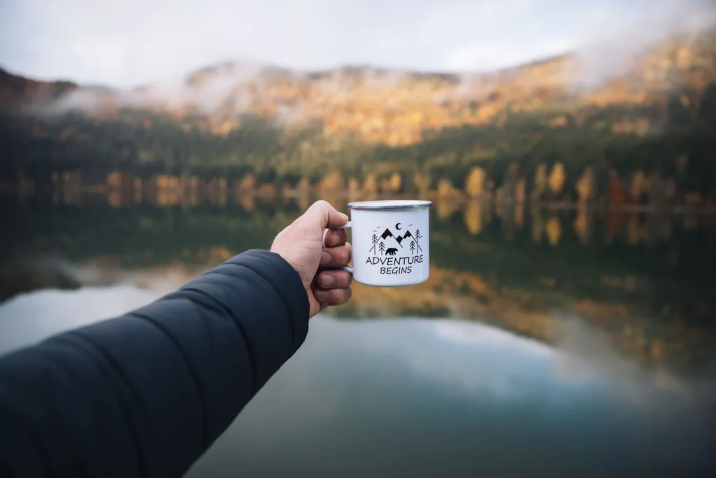 Person holding a cup of coffee that says 'Adventure Begins' on the mug with a lake in the background.