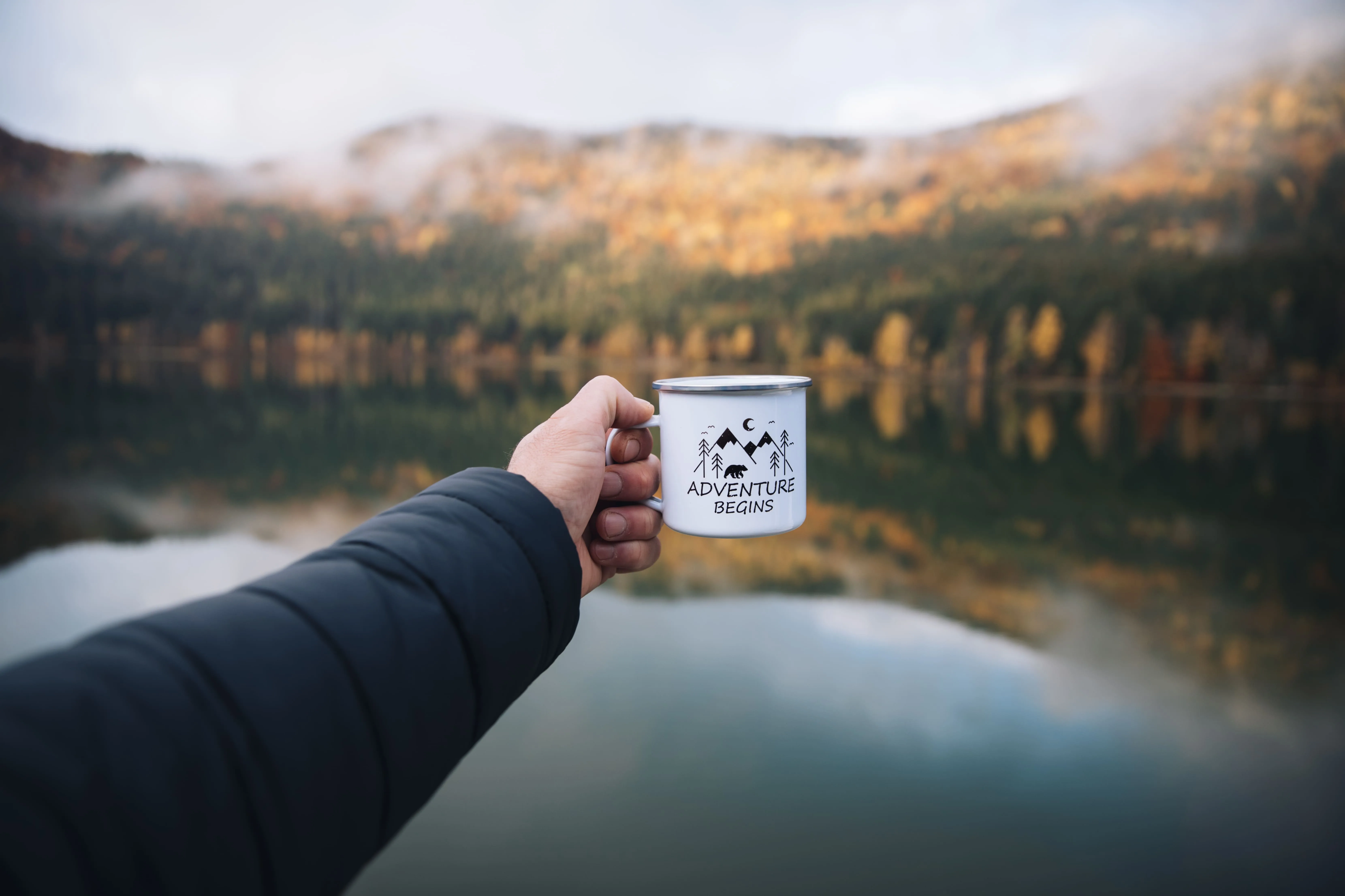 Person holding a cup of coffee that says 'Adventure Begins' on the mug with a lake in the background.