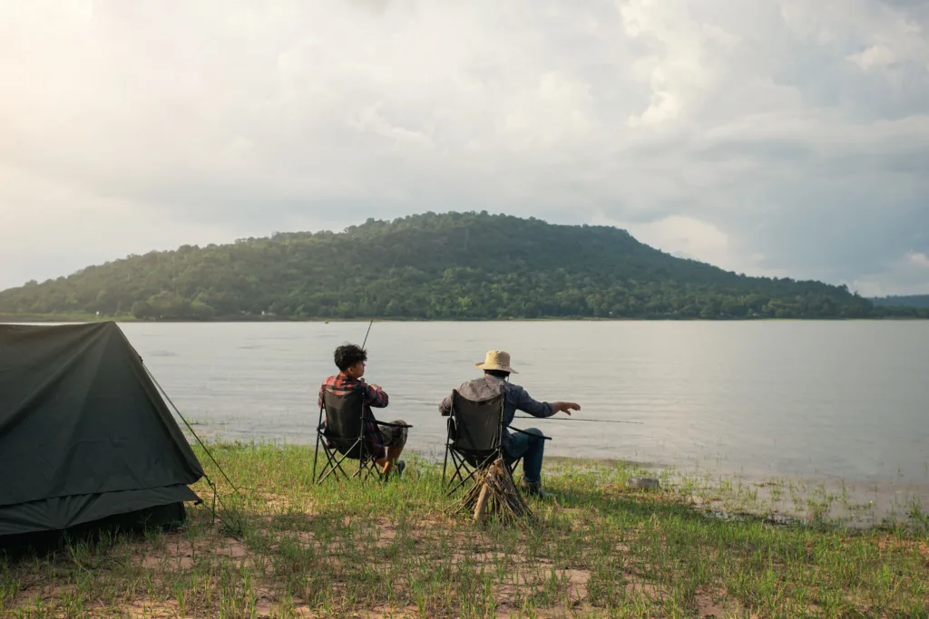 Two friends fishing in a lake with a camping tent in the background.