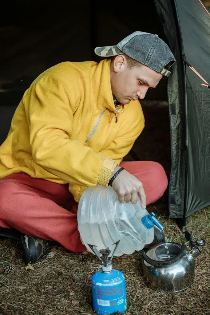 A man pours water into a kettle on a camping stove beside a tent.