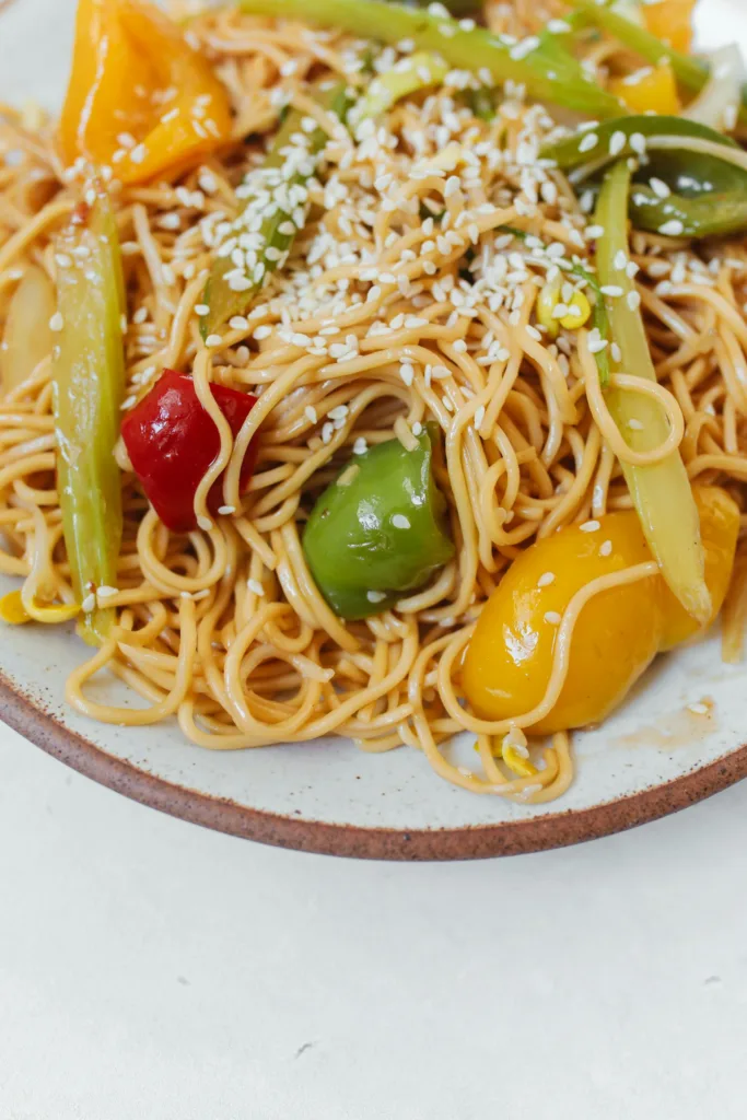 Colorful stir-fried noodles with fresh vegetables and sesame seeds served on a ceramic plate.